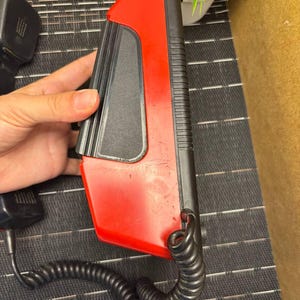 May include: A red and black vintage telephone handset with a coiled cord. The handset has a rectangular shape with a red top and a black base. The cord is black and connects to a black power adapter. The phone is likely from the 1980s or 1990s.