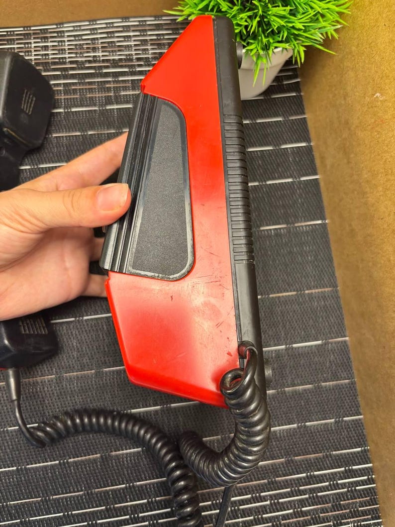 May include: A red and black vintage telephone handset with a coiled cord. The handset has a rectangular shape with a red top and a black base. The cord is black and connects to a black power adapter. The phone is likely from the 1980s or 1990s.