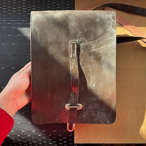 Könnte beinhalten: Eine rechteckige, dunkelbraune Ledertasche mit Klappenverschluss. Die Tasche hat einen langen, dünnen Riemen mit einer Metallschnalle und einem U-förmigen Verschluss. Die Tasche wird von einer Person gehalten.