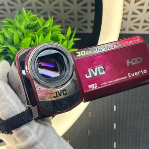 Pode incluir: Uma câmara de vídeo de disco rígido JVC Everio vermelho-escura com lente Konica Minolta, capacidade de armazenamento de 30 GB e modos de gravação híbridos. O dispositivo inclui ranhura para cartão microSD, correia preta e marca em branco.