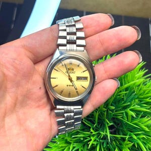May include: A vintage Orient watch with a gold-toned face, silver-colored band, and a day-date display. The watch face features the Orient logo, three stars, and the day of the week and date. The watch is held in a hand.
