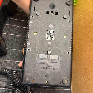 May include: Black vintage telephone base with a coiled cord. The base has a rectangular shape with rounded corners and several small, circular feet. The bottom of the phone has a label that reads "Made in Poland".