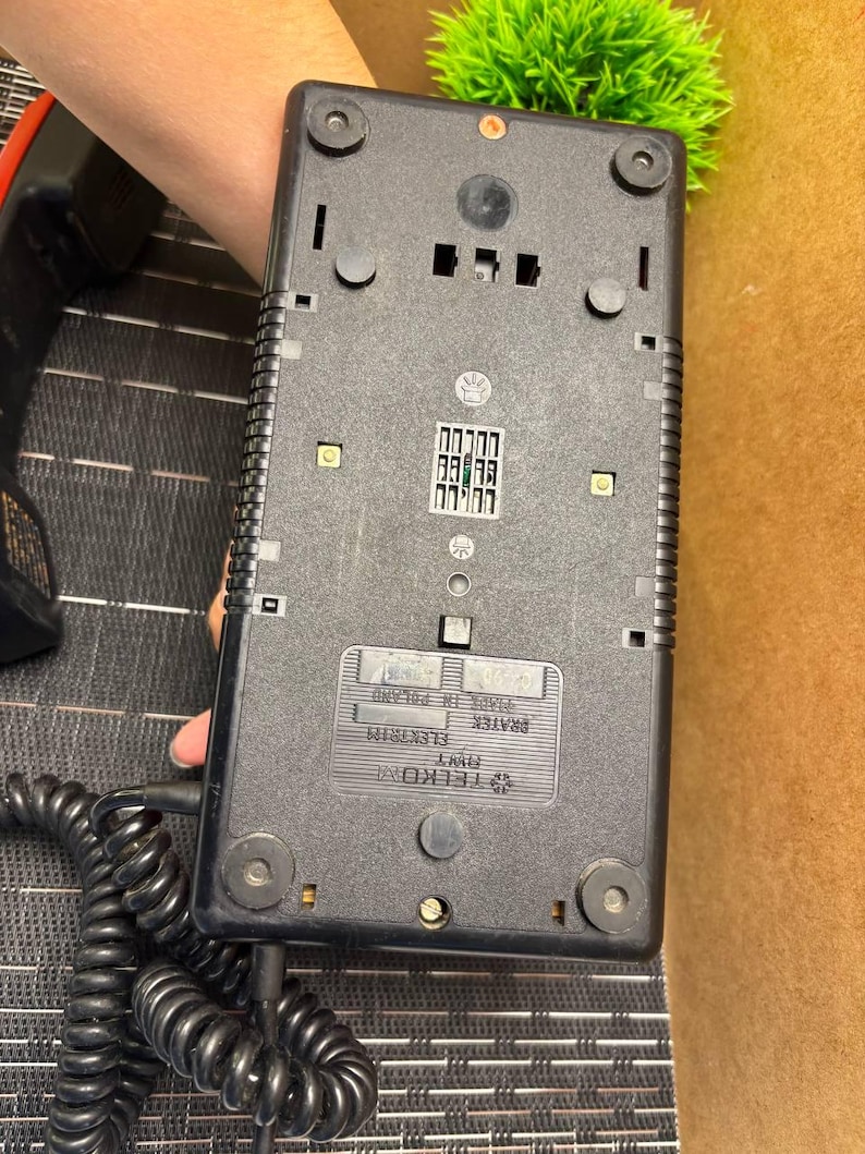 May include: Black vintage telephone base with a coiled cord. The base has a rectangular shape with rounded corners and several small, circular feet. The bottom of the phone has a label that reads "Made in Poland".