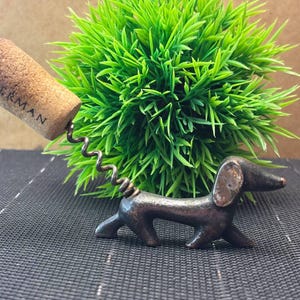 Può includere: Cavatappi a forma di bassotto in metallo marrone. Il cavatappi ha una spirale e un tappo con la scritta "ANKERMAN" stampata. Lo sfondo presenta una pianta verde e una superficie scura e strutturata.