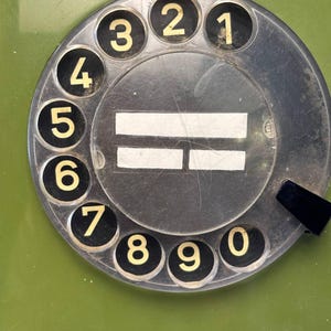 May include: A vintage olive green rotary telephone dial featuring a circular black face with white numbers zero through nine arranged in a clockwise pattern. The center of the dial displays two horizontal white rectangular labels.