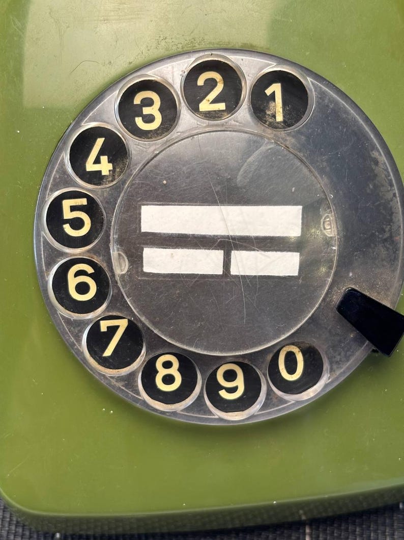 May include: A vintage olive green rotary telephone dial featuring a circular black face with white numbers zero through nine arranged in a clockwise pattern. The center of the dial displays two horizontal white rectangular labels.