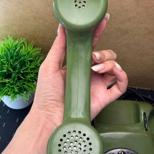 May include: An olive green rotary telephone handset with a circular speaker and microphone section. The handset features a smooth, molded plastic finish with a perforated metal grille on the bottom and a smaller perforated speaker area on the top.