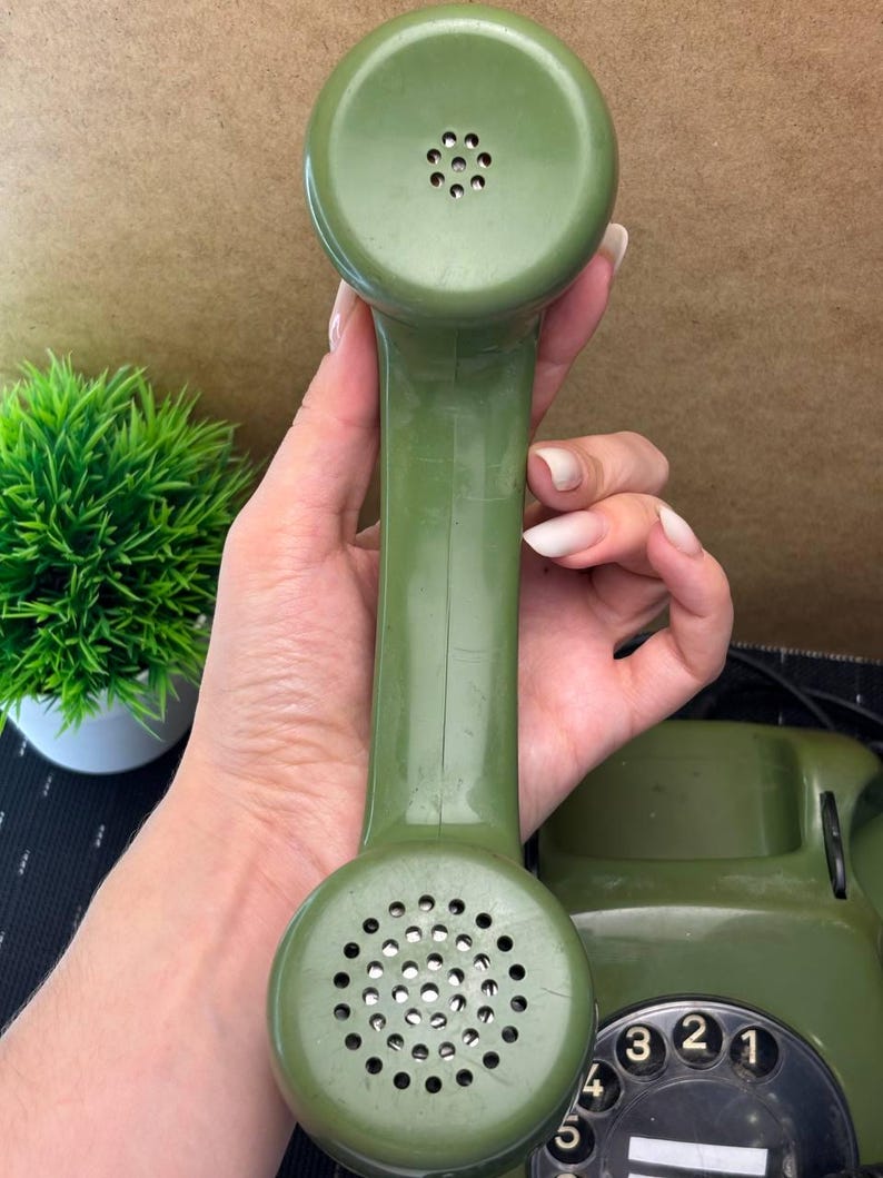 May include: An olive green rotary telephone handset with a circular speaker and microphone section. The handset features a smooth, molded plastic finish with a perforated metal grille on the bottom and a smaller perforated speaker area on the top.