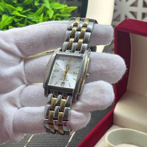 May include: A rectangular wristwatch with a silver and gold-tone metal band and case. The watch face is white with gold-tone hands and markers. The watch is displayed in a red box.