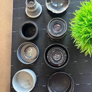 Könnte beinhalten: Eine Sammlung von Vintage-Kameraobjektiven und Zubehör, darunter eine Lupe, Objektivdeckel und verschiedene Objektivkomponenten. Die Artikel sind auf einer dunklen, strukturierten Oberfläche angeordnet, mit einer kleinen grünen Pflanze im Hintergrund.