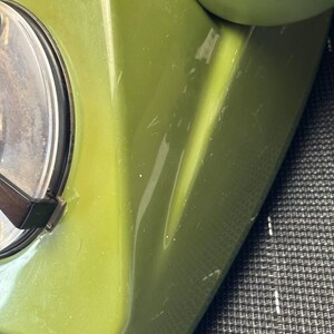 May include: A vintage olive green rotary telephone featuring a smooth, glossy plastic casing. The device includes a circular dial mechanism with a clear plastic cover and a dark brown finger wheel, alongside a matching green handset resting on top.