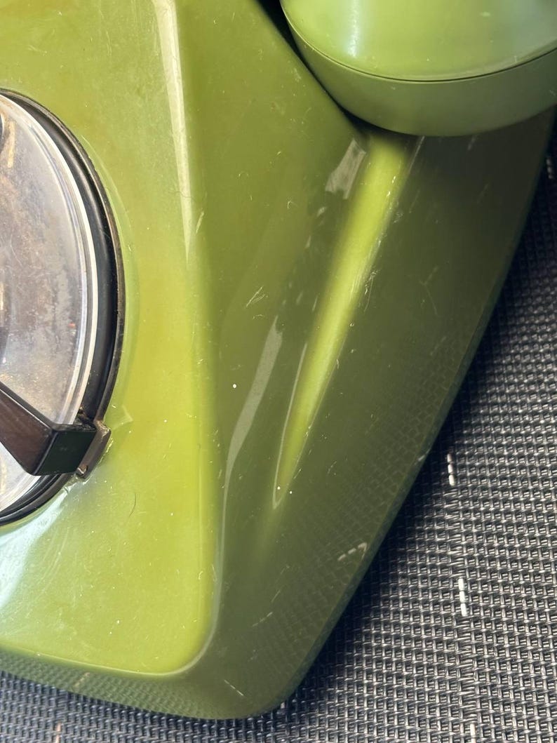 May include: A vintage olive green rotary telephone featuring a smooth, glossy plastic casing. The device includes a circular dial mechanism with a clear plastic cover and a dark brown finger wheel, alongside a matching green handset resting on top.