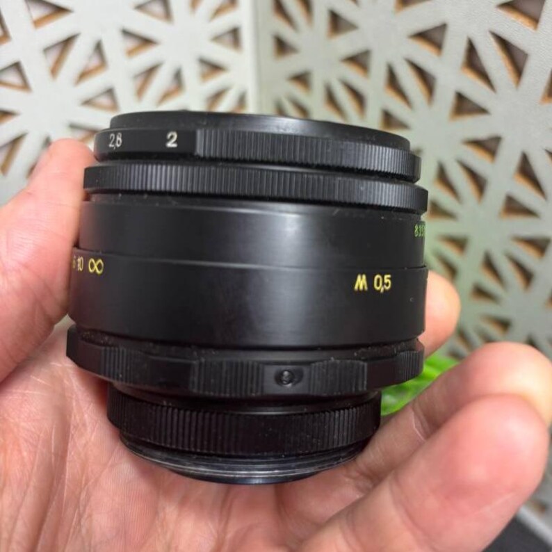 Peut inclure: Un objectif d'appareil photo noir avec des marquages dor&eacute;s, dont des chiffres et des lettres. L'objectif a une bague de mise au point rainur&eacute;e et des r&eacute;glages d'ouverture. L'objectif est tenu dans une main.