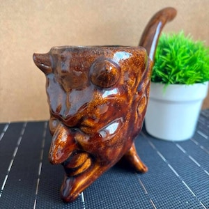 Puede incluir: Una taza de cerámica blanca con un asa negra y el texto "But First, Coffee" en negro, en negrita. La taza está sobre una superficie blanca.