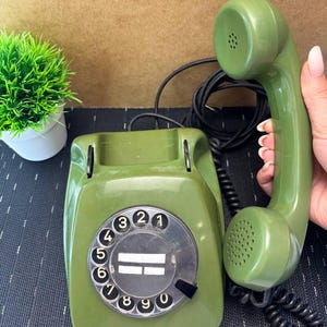 May include: An olive green rotary telephone with a matching handset and a black coiled cord. The circular dial features black numbers from 1 to 0 on a white background, with a central clear plastic label area. The handset is held to the side.