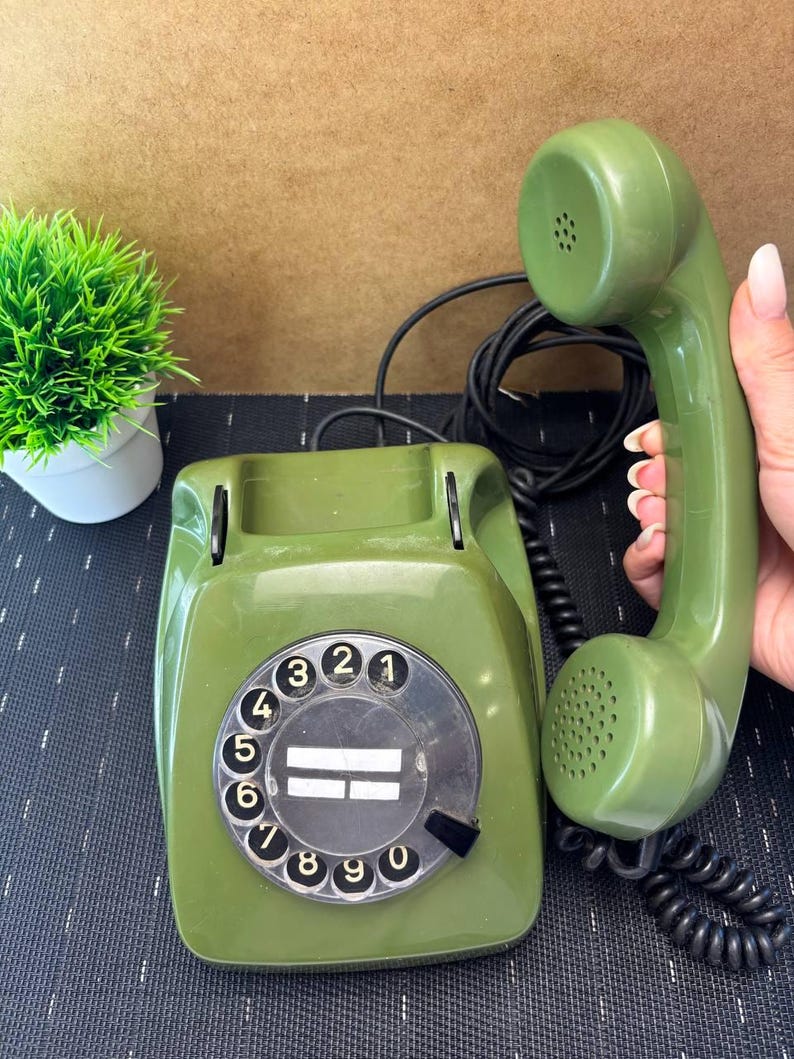 May include: An olive green rotary telephone with a matching handset and a black coiled cord. The circular dial features black numbers from 1 to 0 on a white background, with a central clear plastic label area. The handset is held to the side.
