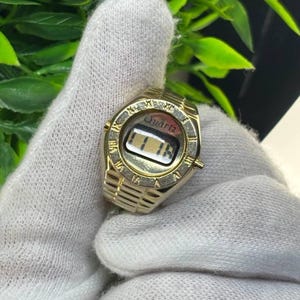 May include: A gold-colored ring watch with a digital display showing the time. The watch face is round with Roman numerals and the word "Quartz" above the display. The ring band has a textured design.