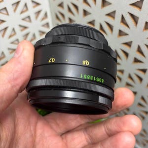 Peut inclure: Un objectif d'appareil photo noir avec des marquages verts et jaunes, tenu dans une main. L'objectif a un design stri&eacute; et les chiffres 0,6 et 0,7 en jaune. Le num&eacute;ro de s&eacute;rie 83513851 est imprim&eacute; en vert.