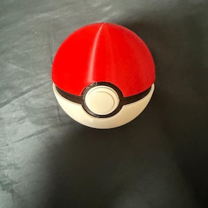 May include: A red and white Poke Ball toy with a black ring around the middle. The toy is made of plastic and has a smooth surface.