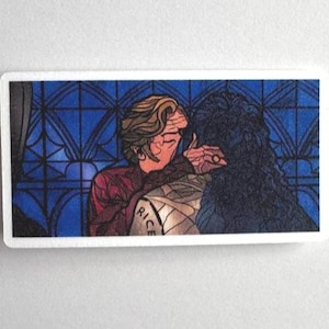 Puede incluir: Una pegatina rectangular con una ilustración de estilo cómic de dos figuras abrazándose. Una figura lleva una camisa roja y la otra tiene el pelo oscuro. El fondo es un diseño de vidriera con paneles azules.