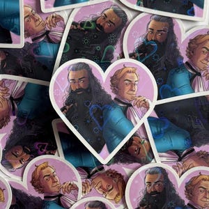 Puede incluir: Pegatinas en forma de corazón con ilustraciones de dos hombres con vestimenta histórica. Uno tiene pelo largo y negro y barba, el otro tiene pelo rubio corto. Las pegatinas tienen un fondo rosa y detalles de corazón azul.