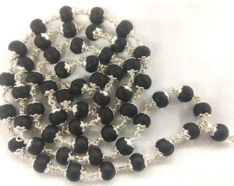 May include: A long necklace featuring black, round beads strung together with silver-colored metal links. The beads have a matte finish, and the metal links have a decorative design. The necklace is displayed on a white surface.