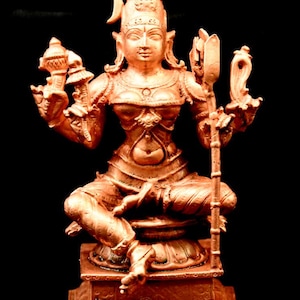 Può includere: Una statua di una divinità indù in rame, seduta con quattro braccia, che tiene uno scettro, un loto e una pentola. La divinità indossa una corona e gioielli decorati. La statua è su una base quadrata.