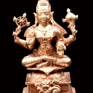 Puede incluir: Una estatua de color bronce de una deidad sentada, adornada con detalles intrincados. La figura sostiene objetos simbólicos en cada mano y está sentada sobre una base de flor de loto. La estatua está sobre un fondo negro.