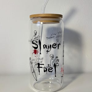 Pode incluir: Um copo de vidro transparente com tampa de bambu e canudo transparente. O copo apresenta ilustrações gráficas pretas e as palavras "Slayer Fuel" em estilo manuscrito. Salpicos vermelhos adicionam uma cor contrastante.