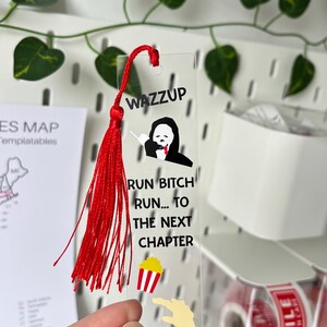 Op de afbeelding: Een doorzichtige, rechthoekige boekenlegger met een rode kwast aan de bovenkant. Het ontwerp bevat de tekst WAZZUP, een afbeelding van een spookachtig figuur, de tekst RUN BITCH RUN... TO THE NEXT CHAPTER, een popcornbakje en een gele hand.
