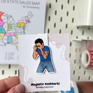 Op de afbeelding: Een magnetische boekenlegger met een illustratie van een persoon met een zonnebril, een blauw t-shirt en een spijkerbroek. De boekenlegger heeft de tekst "Magnetic bookmarks" en de gebruikersnaam van de verkoper. Op de achtergrond is een Amerikaanse staatsverkoopkaart te zien.