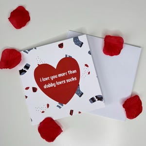 Op de afbeelding: Een wenskaart met een rood hart en de tekst "i love you more than dobby loves socks" is omringd door rode rozenblaadjes. De kaart heeft een witte achtergrond met een patroon van sokken en kleine rode vormen. Een open witte envelop ligt naast de kaart.