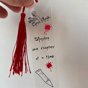 Puede incluir: Marcapáginas transparente con borla roja. El marcapáginas tiene ilustraciones negras de un zombi, salpicaduras de sangre y un lápiz. El texto en el marcapáginas dice "Grrr Argh. Slaying one chapter at a time."