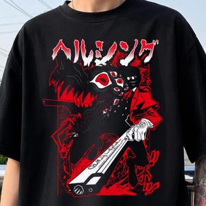 May include: A black t-shirt featuring a graphic print in red, white, and black. The design displays a character in a red suit holding a long firearm, alongside a monstrous creature with multiple eyes. Japanese text appears at the top of the graphic.