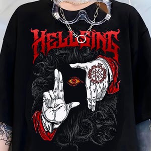 May include: A black t-shirt featuring a red graphic print with the text HELLSING. The design displays two pale hands forming a frame around a single red eye, with a circular red seal on the left hand and dark, swirling abstract patterns.