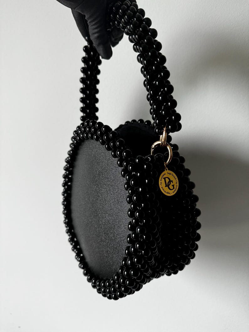 May include: A black round beaded handbag with a black beaded top handle and a gold circular tag with the letters "DG" on it.