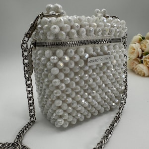 May include: A white beaded crossbody bag with a silver chain strap and a zipper closure. The bag has a small silver tag that reads "DIRECT GLOW."