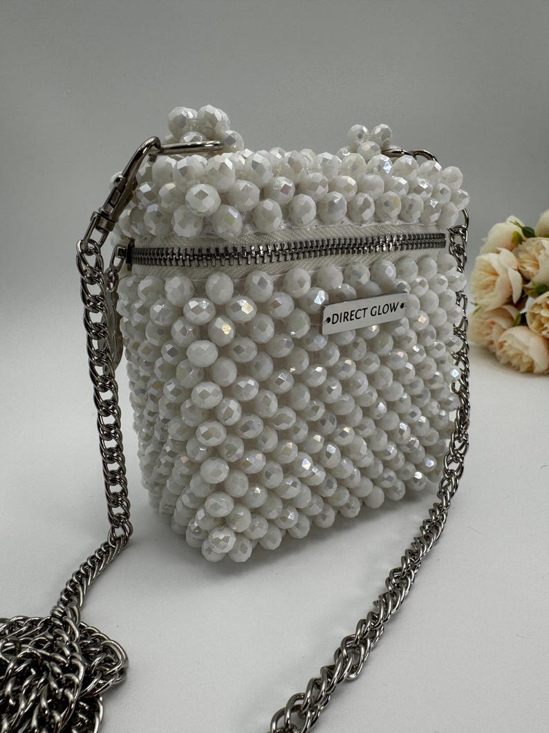 May include: A white beaded crossbody bag with a silver chain strap and a zipper closure. The bag has a small silver tag that reads "DIRECT GLOW."