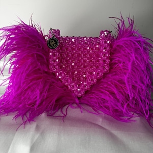 May include: A pink beaded handbag with a heart-shaped design and a small, round, silver charm with the letters "DG" on it. The bag is adorned with a large, fluffy, fuchsia feather trim.