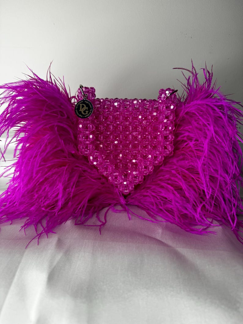May include: A pink beaded handbag with a heart-shaped design and a small, round, silver charm with the letters "DG" on it. The bag is adorned with a large, fluffy, fuchsia feather trim.
