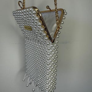 May include: A white pearl beaded clutch bag with a gold chain strap. The bag is open and the interior is lined with a white fabric. The bag has a fringe detail at the bottom.
