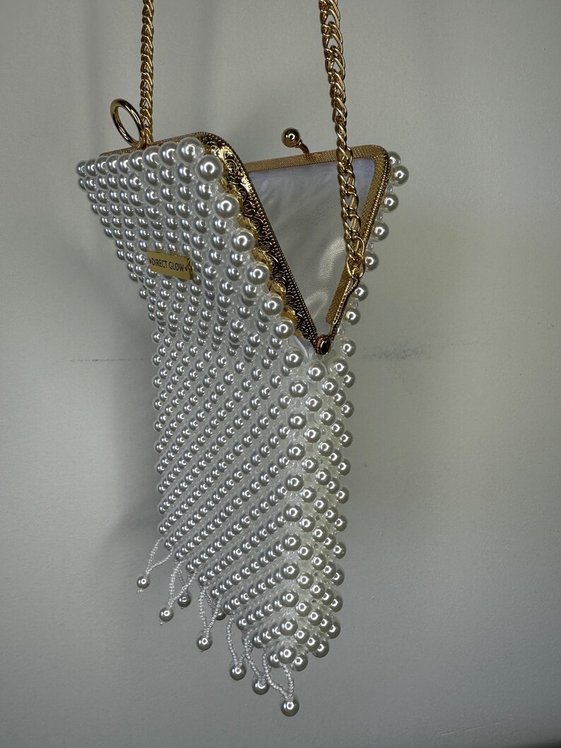 May include: A white pearl beaded clutch bag with a gold chain strap. The bag is open and the interior is lined with a white fabric. The bag has a fringe detail at the bottom.
