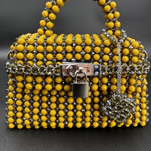 May include: A small yellow and gray beaded handbag with a silver lock and chain. The bag has a top handle and a detachable charm.
