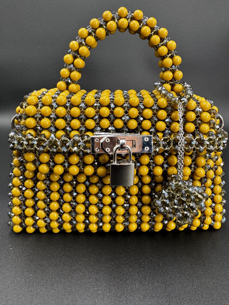 May include: A small yellow and gray beaded handbag with a silver lock and chain. The bag has a top handle and a detachable charm.