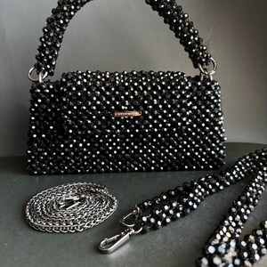 May include: A black beaded handbag with a top handle and a detachable chain strap. The bag has a rectangular shape and is made of small, black beads. The brand name "DIRECT GOLD" is visible on the front of the bag.