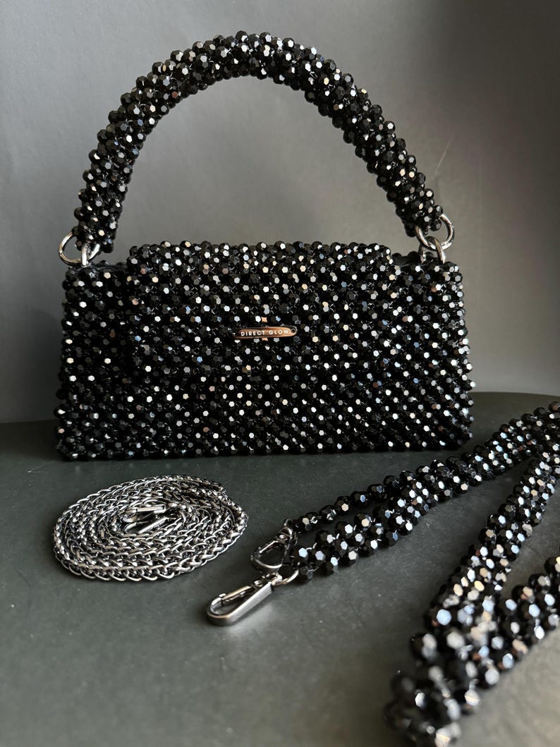May include: A black beaded handbag with a top handle and a detachable chain strap. The bag has a rectangular shape and is made of small, black beads. The brand name "DIRECT GOLD" is visible on the front of the bag.
