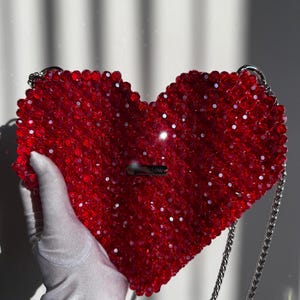 May include: A red heart-shaped beaded bag with a silver chain strap. The bag has a small silver tag that says "#GLOW".