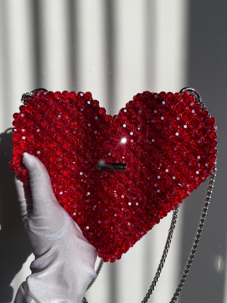 May include: A red heart-shaped beaded bag with a silver chain strap. The bag has a small silver tag that says "#GLOW".