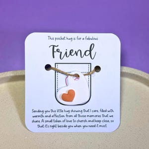 Könnte beinhalten: Eine weiße Karte mit dem Text "This pocket hug is for a fabulous Friend". Ein kleines weißes Herz mit einem orangefarbenen Herz ist an der Karte befestigt. Die Karte enthält eine Botschaft über Freundschaft.
