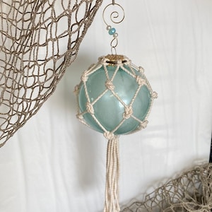 Op de afbeelding: Een blauwe glazen drijver ornament met een wit touwnet en een witte kwast. De ornament hangt aan een gouden draadlus met blauwe en witte kralen.